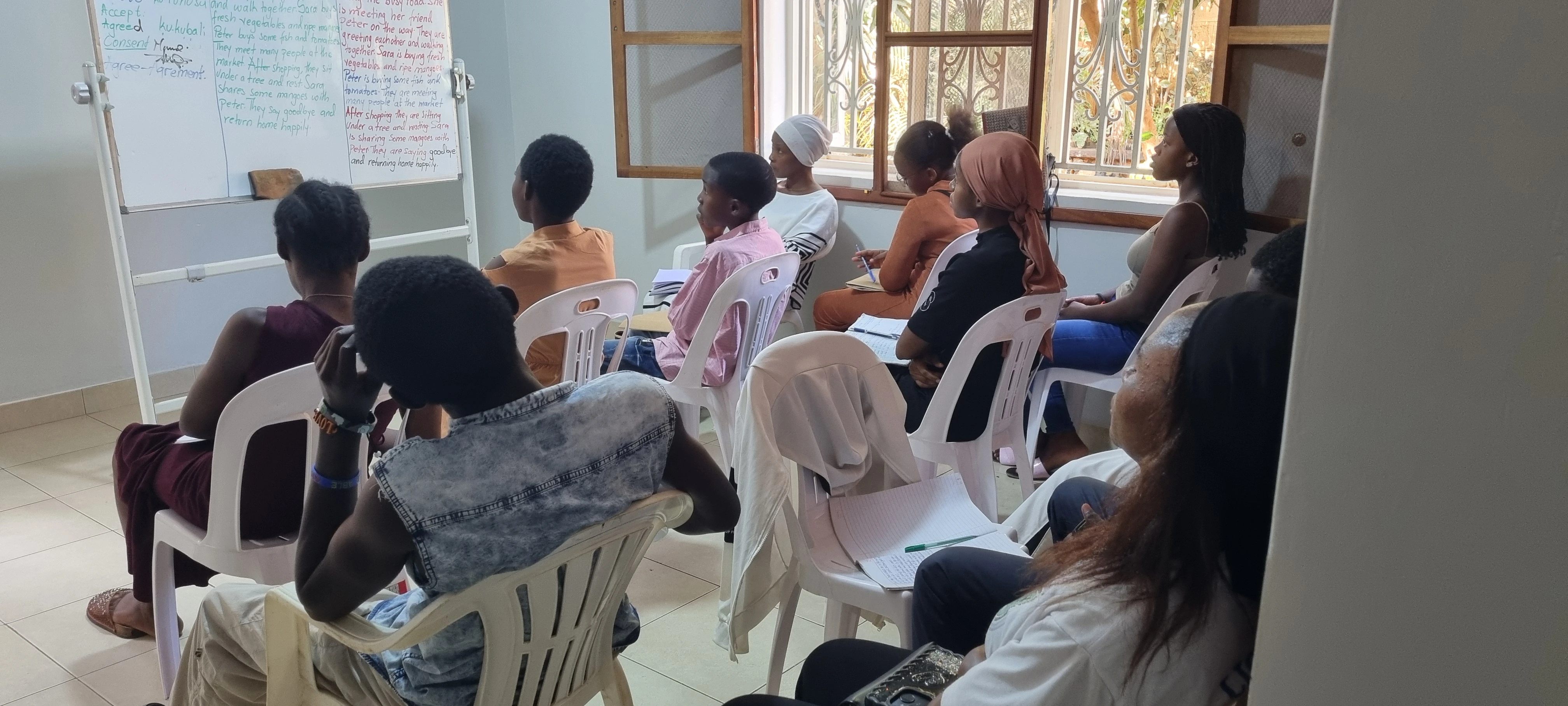 YAREN English learners seated in rows of white plastic chairs facing a whiteboard covered in handwritten notes
